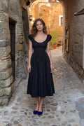 Front view of woman wearing black square neck smocked midi dress in rustic alley