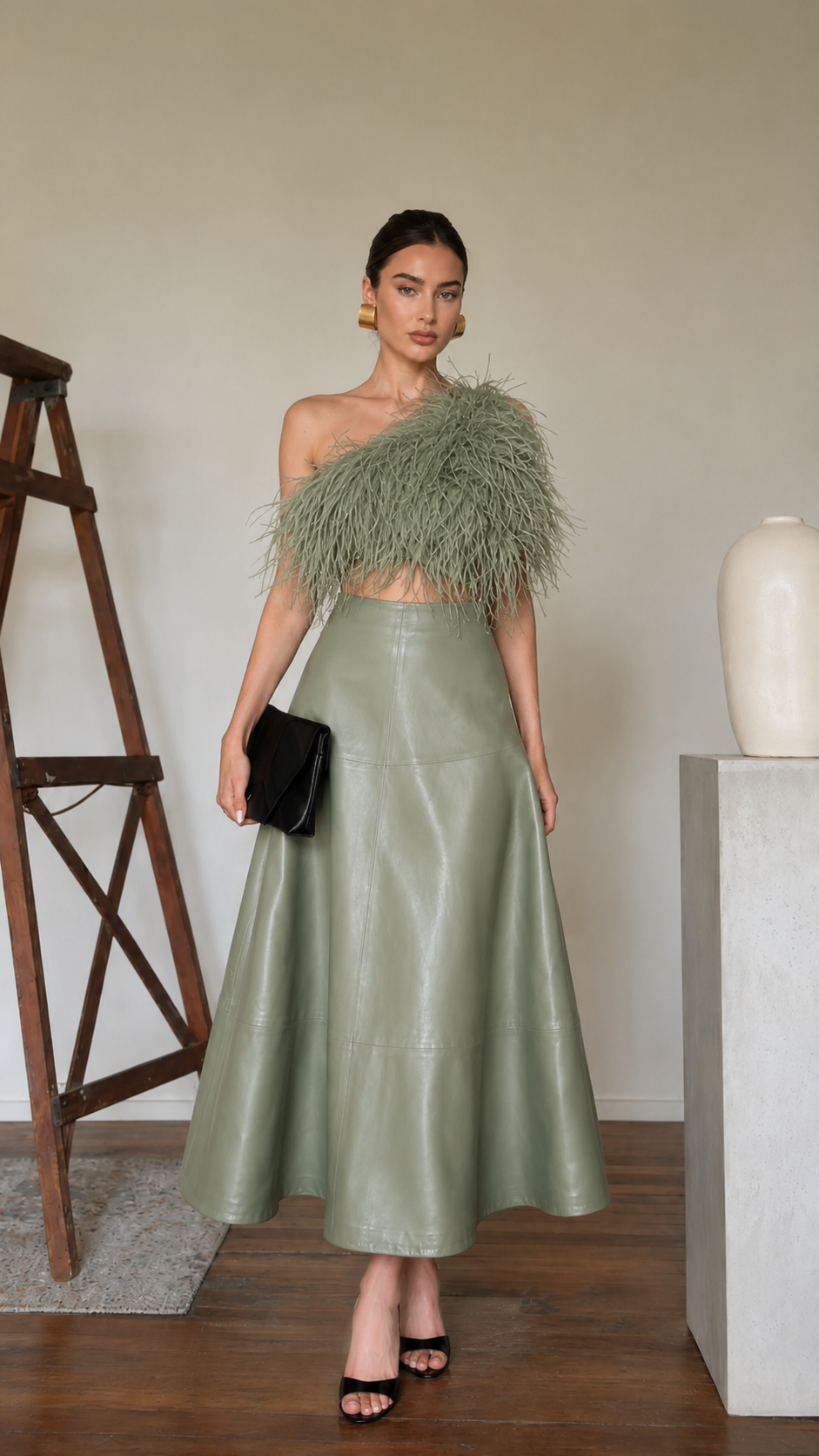 Model wearing a sage high-waisted faux leather A-line midi skirt with a one-shoulder feather top